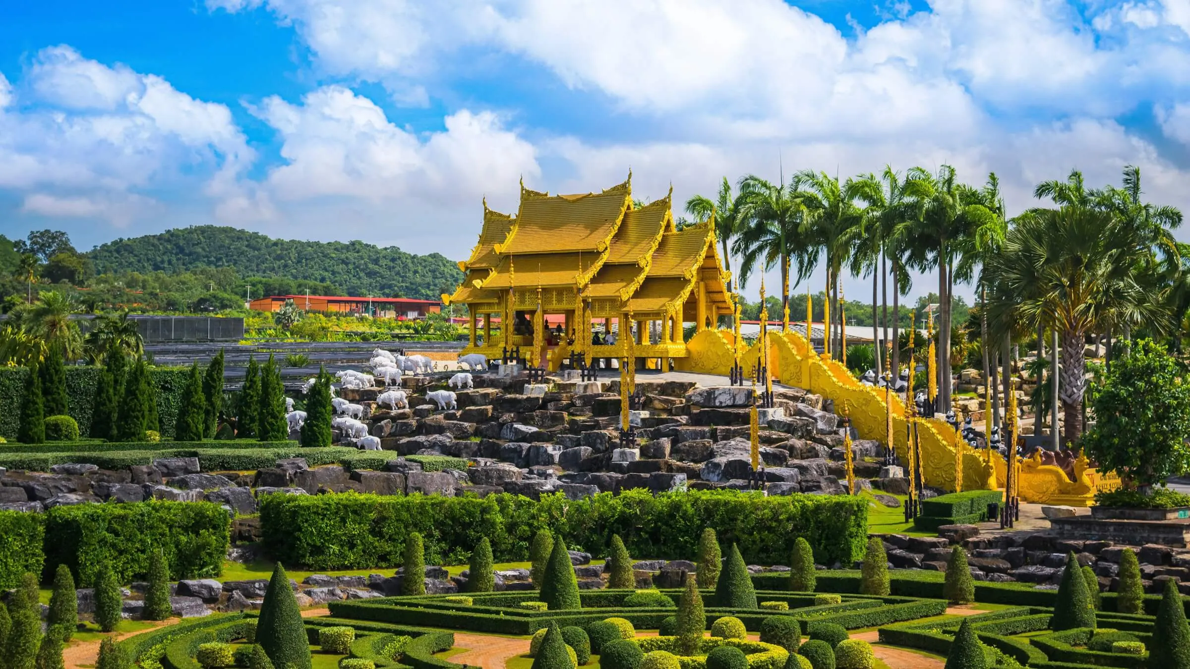 A golden Thai-style pavilion on a rock garden with white elephant statues, palms behind it and rows of clipped topiary cones in front, at Nong Nooch Tropical Garden